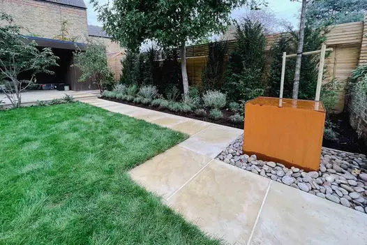 Corten steel water tank sits on large pebbles next to wide path of Harvest sawn sandstone paving with 2 right-angled bends.