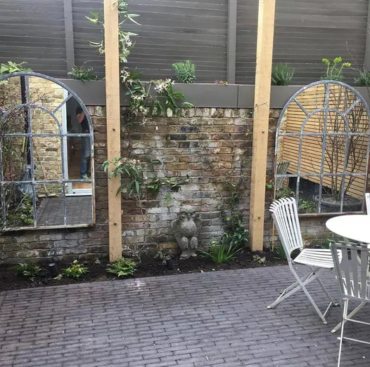 Small garden showcasing two window-style mirrors on a brick wall with climbing plants, and metal furniture set on an Amersham clay paver patio.