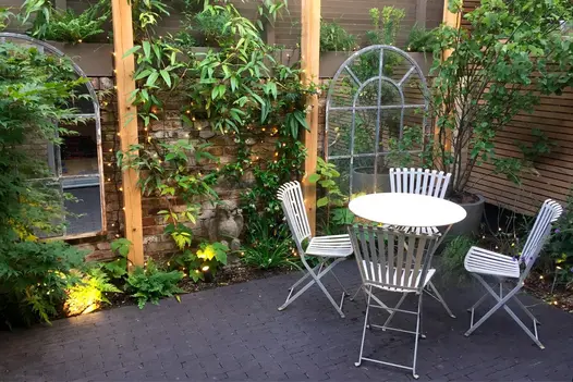 Small garden with 2 window style mirrors with plants growing up the brick wall they hang from, metal furniture sits on amersham clay paver patio.