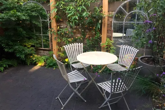 Small garden with two window-style mirrors on a brick wall adorned with climbing plants, and metal furniture placed on an Amersham clay paver patio.
