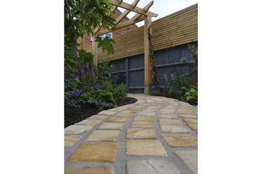 Low view of curvy path using Mint sandstone setts runs through wooden pergola with planting beds either side of the cobble path.
