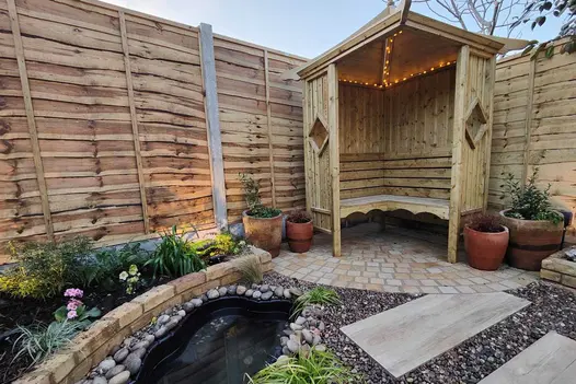Covered wooden bench in corner of fenced garden, sits on Mint Sandstone Setts next to brick raised bed and porcelain paving.