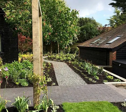 Between newly planted beds gravel path leads away from path of Lucca Dutch clay pavers and turns corner behind brick building.