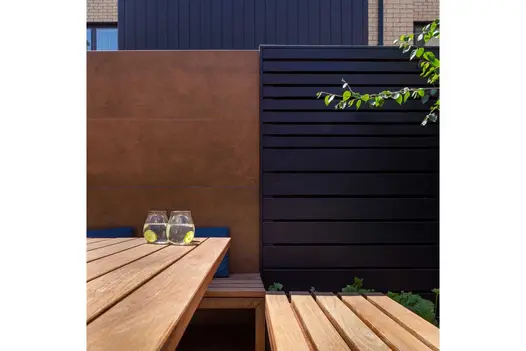 View down length of wooden table and bench seat to fence of a dark panel next to Steel Corten external cladding. Built by Nordland.