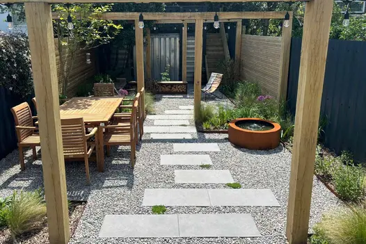 Slabs of Steel Grey Porcelain Paving with pebbles around create stepping stones through pergola, water feature and dining table either side.
