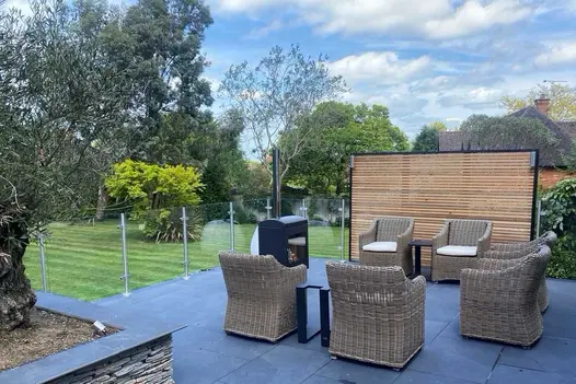 Patio of Florence dark porcelain outdoor tiles UK with olive tree, 6 rattan armchairs and wood burner overlooks lawn and trees.