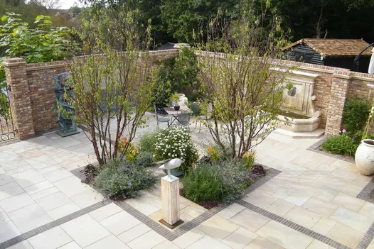 Large square walled area of Harvest sawn sandstone paving divided into quadrants by clay paving stripes. Planted beds in centre.