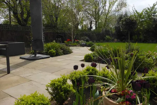 Raj Green Indian Sandstone patio paving runs through planting beds and out onto large lawn in fenced in garden.