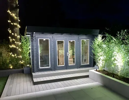 Outdoor building and decking area using garden lighting shows pebble grey brushed composite decking as small patio and steps up.
