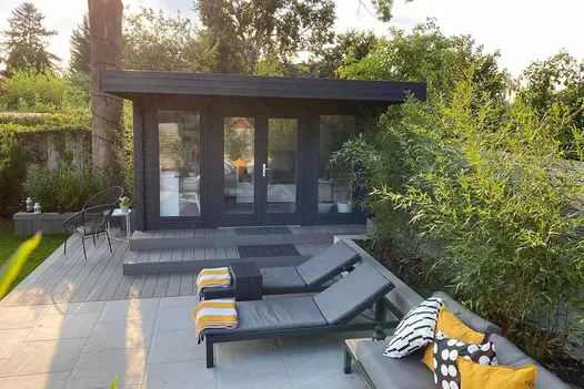Sunloungers and bistro set sit on porcelain paving and Pebble Grey Brushed composite decking in front of glass-fronted garden room.