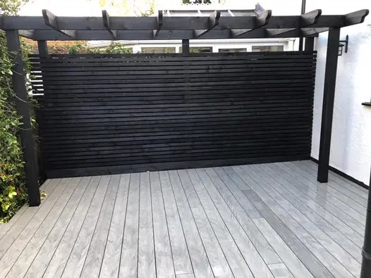 Black slatted fence sits next to black wooden pergola with luna designboard composite decking running underneath.