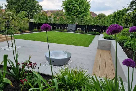Square lawn sits in the middle of polished concrete porcelain patio with black slate water feature, flowerbeds surround.