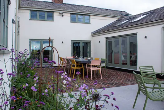 White clad house in the distance, polvere porcelain paving slabs used next to brown clay pavers with purple plants in the foreground.