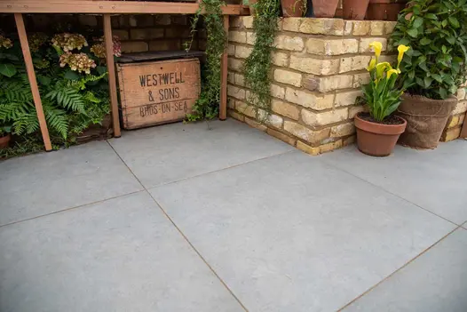 Wooden crate, hydrangea and ferns sit under staging on Anthracite Porcelain paving next to brick wall and planted terracotta pots.