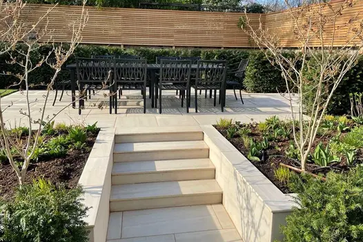 In walled garden, Faro Porcelain pencil round steps rise to patio with dining set and trimmed hedge. Design by Shoots and Leaves.