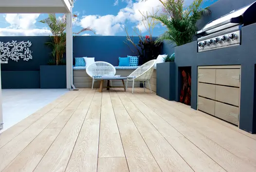 Blue rendered walls surround outdoor cooking area with limed oak millboard decking hosting seating area, white pergola to the left.