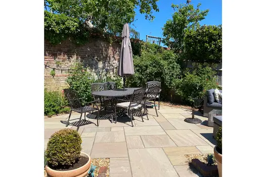 Dining set on Raj Green Indian sandstone patio with brick wall and tall green bushes behind and flowerpots in front.