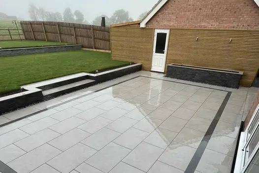 Batten fence, raised beds and water feature disguise tall brick wall on border of oblong Florence Grey patio at back of house.