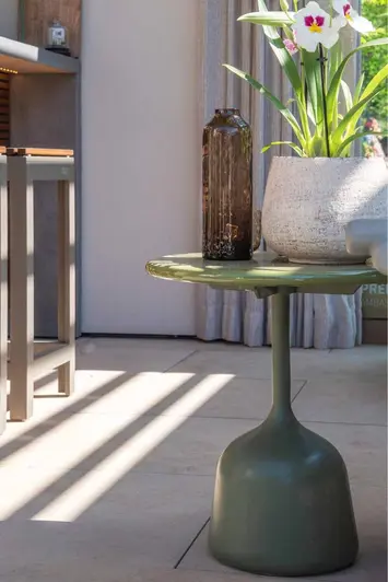 Arty shot of table that looks like an upside down wine glass, with vase and plant pot on top, sits on Jura Beige porcelain paving at RHS Chelsea.
