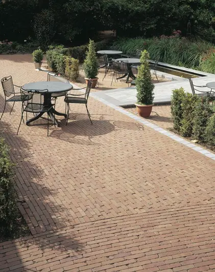 Three sets of metal garden furniture sit on roma brick paving patio with ramp leading up to long narrow pond feature.