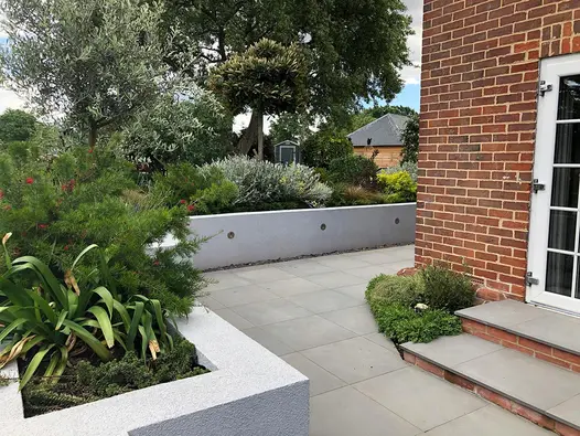 Steel Grey porcelain patio wrapping around the side of a red brick house with a rendered retaining wall to house raised planted beds.