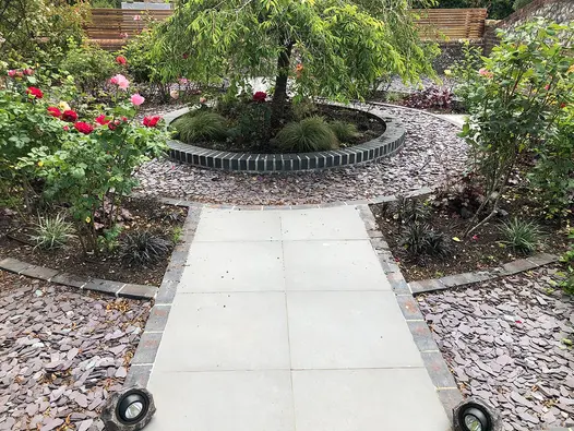 Steel Grey porcelain path leading to a circular brick bed with a tree as the centre piece and surrounding rings of slate chipping and flower beds.