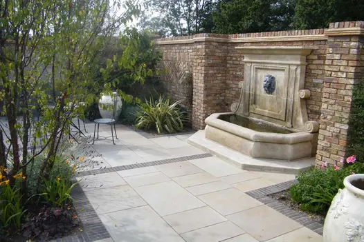 Large water trough with lion head spouting water above it, against wall in area paved with Harvest sawn sandstone slabs.