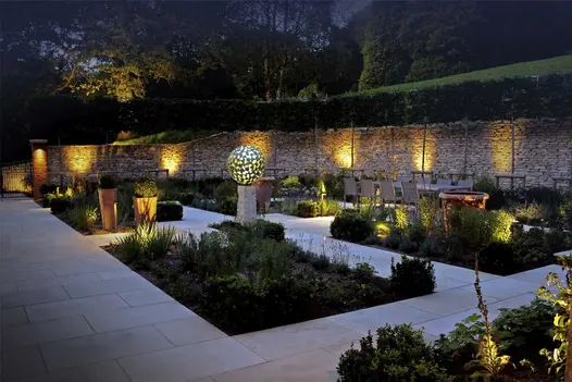 Pierced ball on pillar in centre of 4 oblong beds. Paths of Harvest sawn sandstone paving. Illuminated wall in background. 
