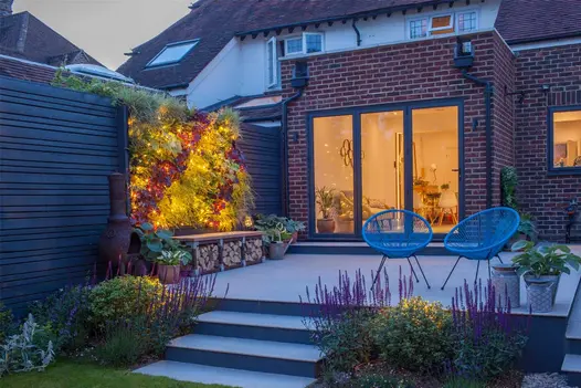 3 steps rise to square patio of Porcelain Grey outdoor tiles outside room with bifold doors. Log storage bench and 2 blue chairs.
