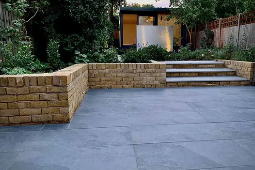 Wide Slab Coke porcelain steps rise between low brick flanking walls from matching paved area to path between planted borders.