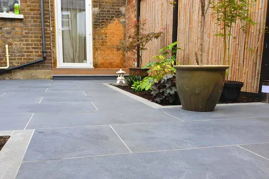 Area outside back door paved with Slab Coke outdoor tiles by Crocus Landscapes. Narrow grey porcelain tiles edge rectangular bed.
