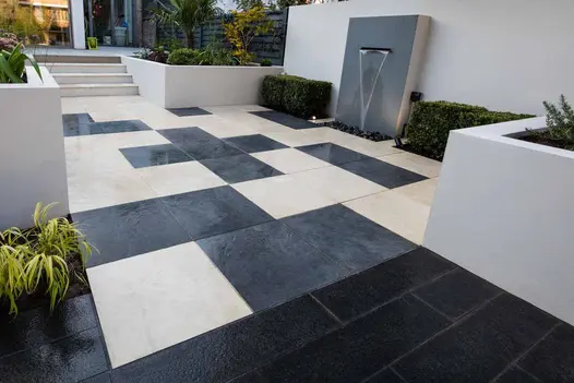 Steps down to Khaki and Slab Coke porcelain paving laid in random geometric pattern, with wall water feature between low hedges.