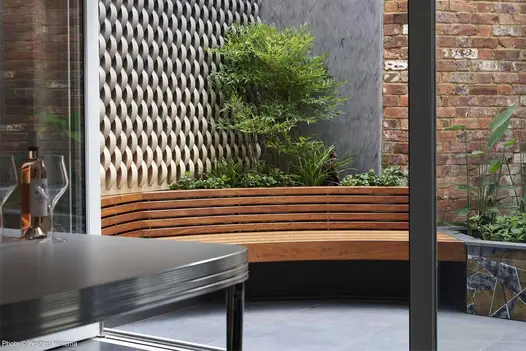 Slab Coke porcelain paved corner of courtyard, with triangular raised bed behind wooden bench. Design by Rosemary Coldstream.
