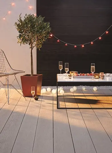 Wire chair and glass table with champagne glasses sit on smoked oak millboard decking, with potted plant and fairy lights.