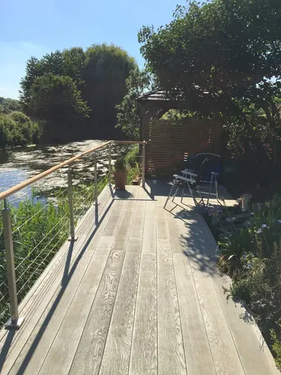 Metal chairs on sunny terrace of Smoked Oak Millboard decking, with steel-wire railings overlooking river. Design by LandArt.