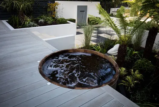 Wooden barrel filled with water built into smoked oak millboard decking with palm trees and white rendered walls.