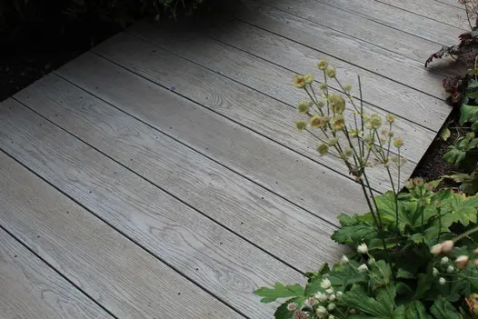 On edge of planted border, close view of path of Millboard Smoked Oak decking boards all of equal length, laid across pathway.