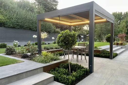 Lit up proteus grey pergola sits on patio with lawn and flowerbeds clad with steel corten porcelain cladding surrounding.