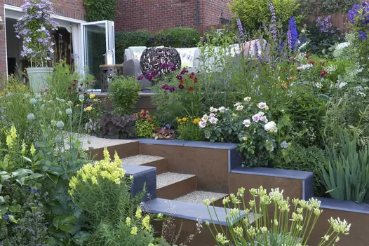 Lots of wild flowers in multi level garden show pebble dashed steps leading down with steel corten designclad lining the steps and walls.