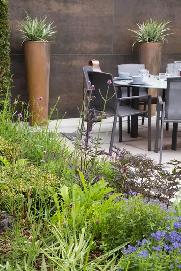 Thickly planted bed next to dining set. 2 tall planted pots stand against wall faced with Steel Dark porcelain cladding panels.