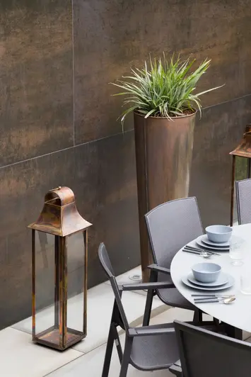 Tall empty copper and glass lantern and tall pot stand against wall faced with Steel Dark DesignClad panels, behind dining set.