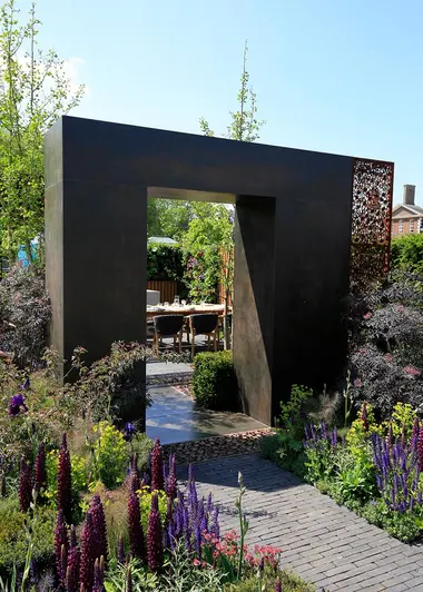 Chunky square arch faced with Steel Dark external cladding straddles path leading between planted beds to seating area.