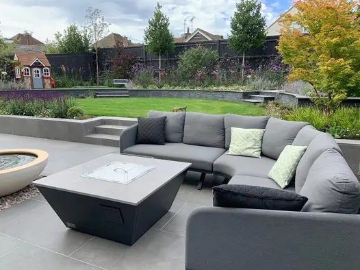 Large outdoor curved sofa around a firepit sitting on a Steel Grey porcelain patio and in front of low retaining wall leading to a raised lawn.