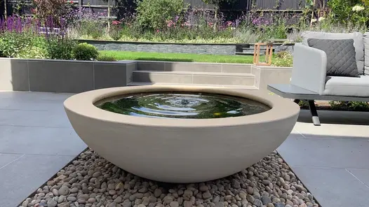 Large stone bowl water feature sitting on a bed of smooth pebbles and surrounded on all sides by Steel Grey porcelain paving.