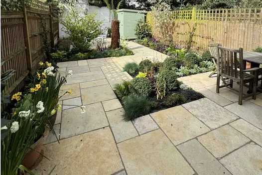 Daffodils planted in pots line a fence with dining table on the other side. Antique Yellow Limestone Paving used with flowerbeds cut out.