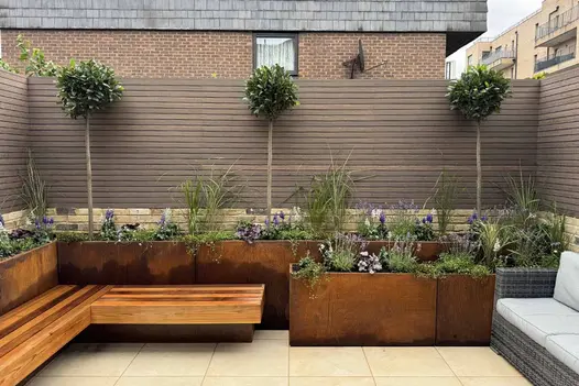 View from middle of garden of corten steel tall trough planters, filled with green and purple plants, framed by chestnut-colored composite battens.