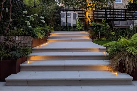 Spoltlights line Faro Porcelain 36mm Bullnose Steps in a garden leading to a tree surrounded by garden lighting.