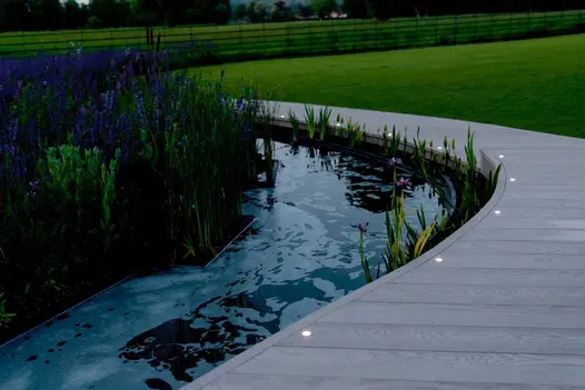 Dusk views show smoked oak Millboard curving around a pond with built-in flowerbeds, with spotlights lining the edge of the decking.