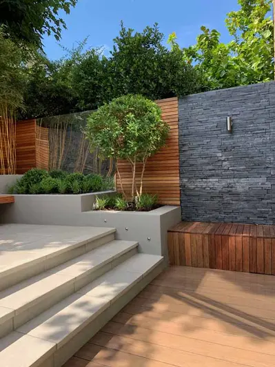 Cream steps lead away from Traditional DesignBoard decking area with stone clad walls and raised planter beds surrounding.
