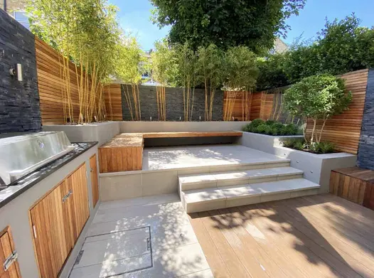 Cream steps lead down to a Traditional DesignBoard decking area, framed by stone-clad walls and raised planter beds, with a BBQ area to the left.